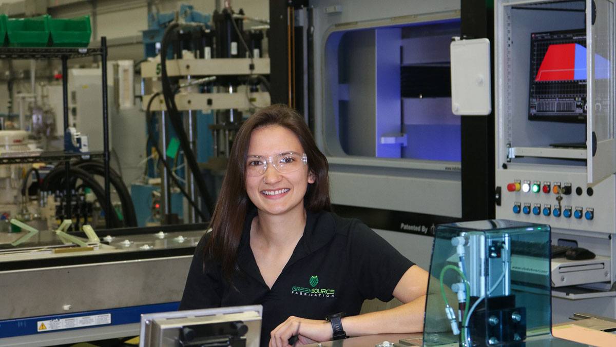 A woman wearing safety glasses smiles at the camera in a lab with industrial machinery.