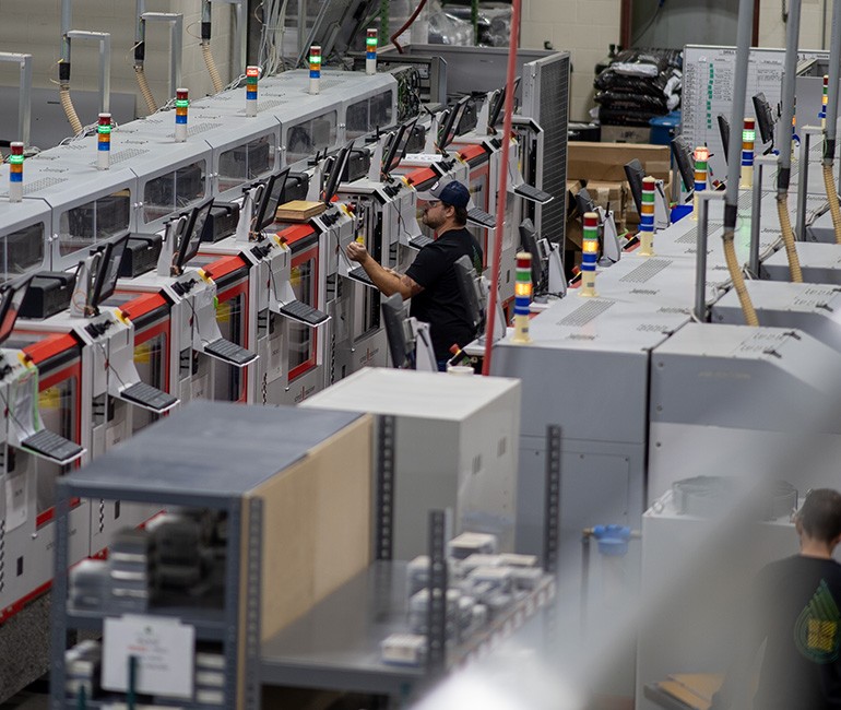 A worker operates a row of industrial machines with control panels and indicator lights in a manufacturing facility.