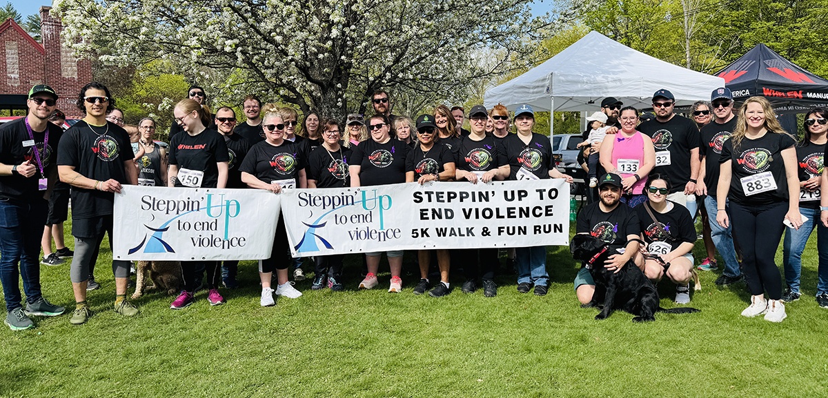 A large group of people pose outdoors on grass holding banners for "Steppin' Up to End Violence 5K Walk & Fun Run," with trees and tents in the background.